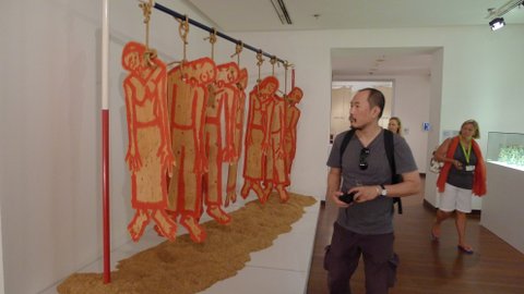 Vasan Sitthiket highlighting the plight of Thai farmers through an installation Committing Suicide Culture: The Only Way Thai Farmers Escape Debt (1995), featuring wooden cutouts of farmers hanging by the noose on a pole painted in Red white and blue, the Thai National colours, hovering over mounds of rice husks. Fra utstillingen Negotiating Home, History, Nation.